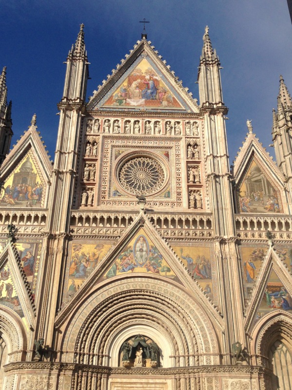 The ornate Gothic facade of the Duomo di Orvieto against a blue sky, with golden mosaics and rose window